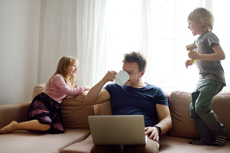 Man working while sitting on sofa with children playing beside him