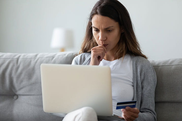 A woman with a worried look on her face, holding her credit card in front of a laptop.